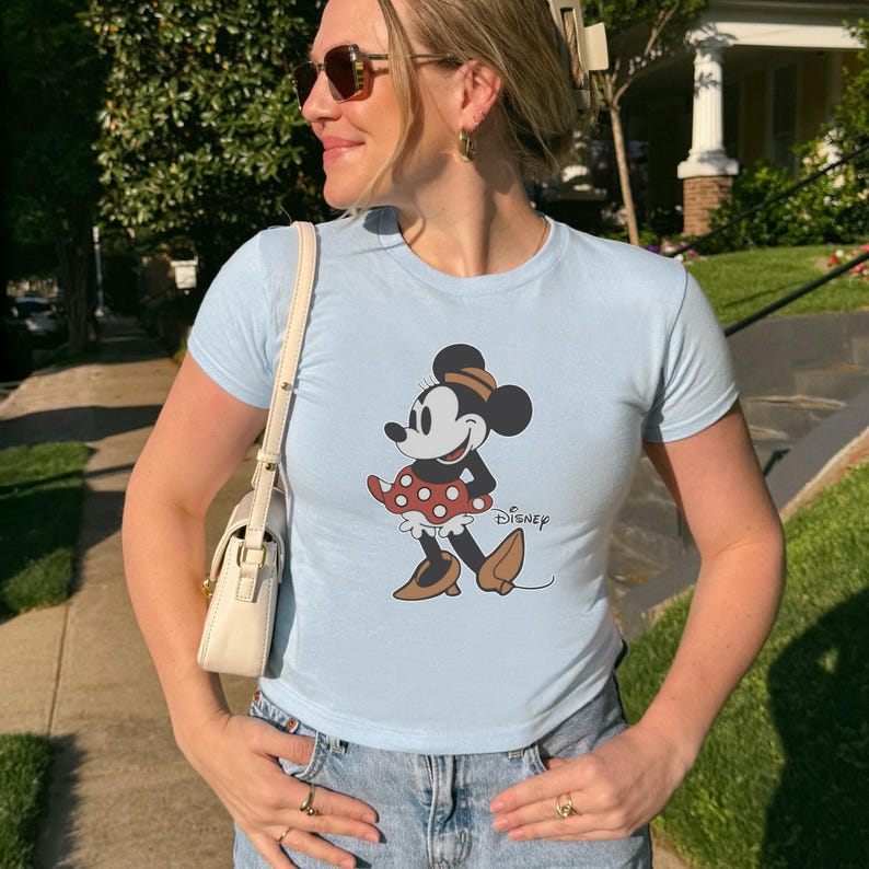 May include: Light blue t-shirt featuring a Minnie Mouse graphic. The cartoon character is depicted in a red and white polka dot dress, brown shoes, and a matching hat. The word "Disney" is printed below the graphic.