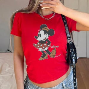 May include: A red t-shirt featuring a graphic of Minnie Mouse in a polka-dot skirt and brown shoes. The Disney logo is printed below the character. The shirt has short sleeves.