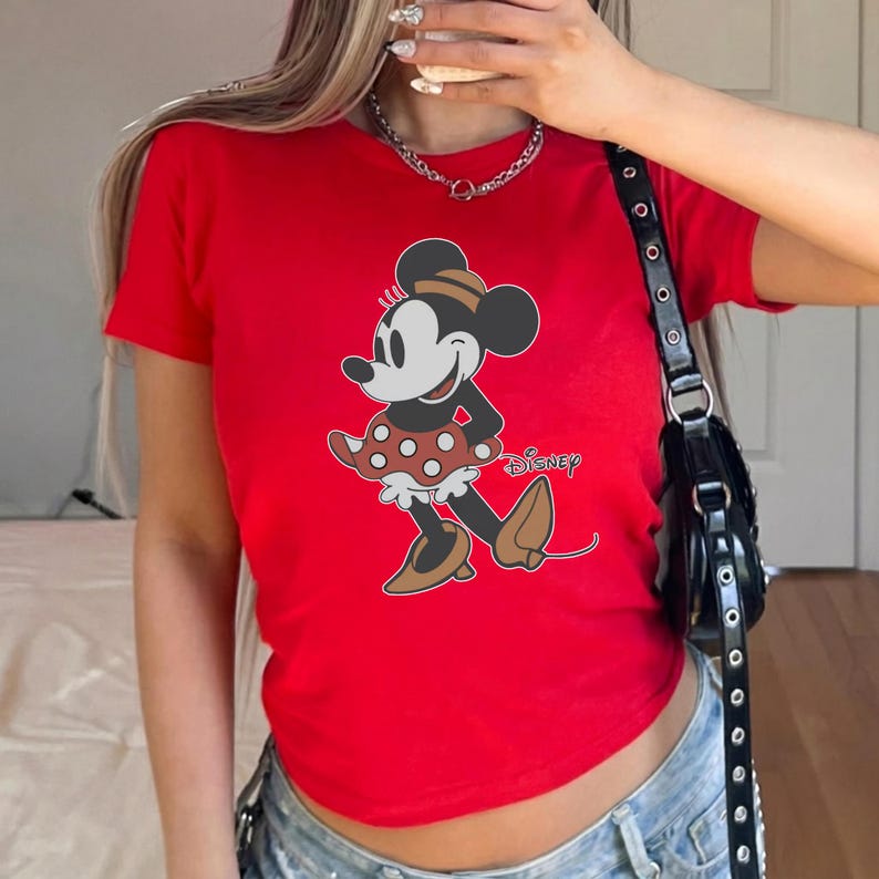 May include: A red t-shirt featuring a graphic of Minnie Mouse in a polka-dot skirt and brown shoes. The Disney logo is printed below the character. The shirt has short sleeves.