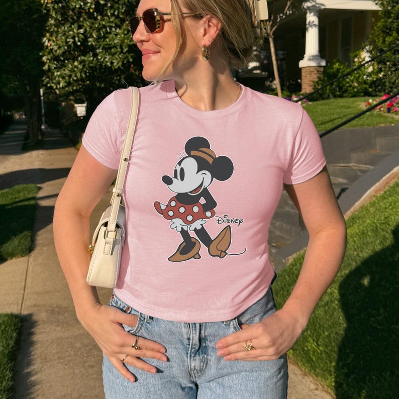 May include: A light pink t-shirt featuring a Minnie Mouse graphic. The cartoon character is depicted in a classic pose, wearing a red polka-dot skirt, brown shoes, and a matching hat. The Disney logo is printed below the graphic.