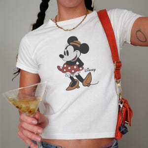 May include: White t-shirt featuring a classic Minnie Mouse graphic. The cartoon character is depicted in a red polka-dot dress and brown shoes. The word "Disney" is printed below the character. A gold necklace and a red purse with a gold chain are also visible.