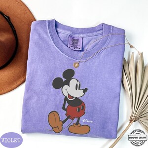 May include: A violet Comfort Colors t-shirt with a Mickey Mouse graphic. The shirt has a crew neck and short sleeves. A gold necklace with a pendant is on the shirt. The word "Disney" is printed on the shirt.