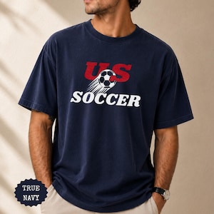 Op de afbeelding: Marineblauwe t-shirt met de tekst "US SOCCER" in rood en wit. Het ontwerp bevat een voetbal met bewegingslijnen. Het shirt heeft een relaxte pasvorm.
