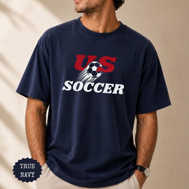 Op de afbeelding: Marineblauwe t-shirt met de tekst "US SOCCER" in rood en wit. Het ontwerp bevat een voetbal met bewegingslijnen. Het shirt heeft een relaxte pasvorm.
