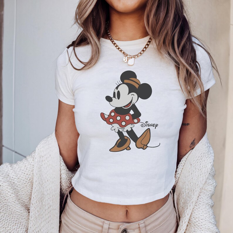 May include: White crop top featuring a graphic of Minnie Mouse in a red polka dot skirt and brown shoes. The Disney logo is printed on the shirt. The shirt has short sleeves and a crew neck.
