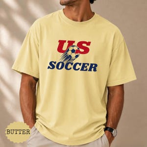Op de afbeelding: Boterkleurig T-shirt met de tekst "US SOCCER" in rood en blauw. Het ontwerp bevat een voetbal met bewegingslijnen. Het shirt is een casual stijl met korte mouwen.