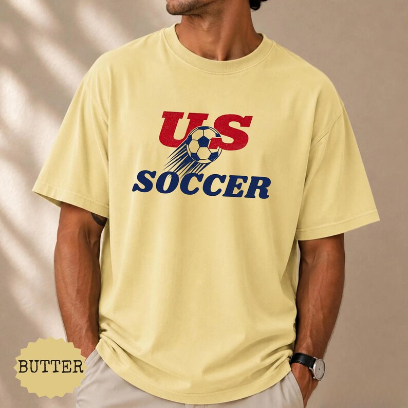 Op de afbeelding: Boterkleurig T-shirt met de tekst "US SOCCER" in rood en blauw. Het ontwerp bevat een voetbal met bewegingslijnen. Het shirt is een casual stijl met korte mouwen.