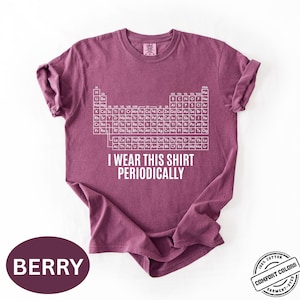Może przedstawiać: Koszulka w kolorze jagodowym z białym wzorem układu okresowego i napisem "I WEAR THIS SHIRT PERIODICALLY". Koszulka wykonana w 100% z bawełny i farbowana w kawałku.