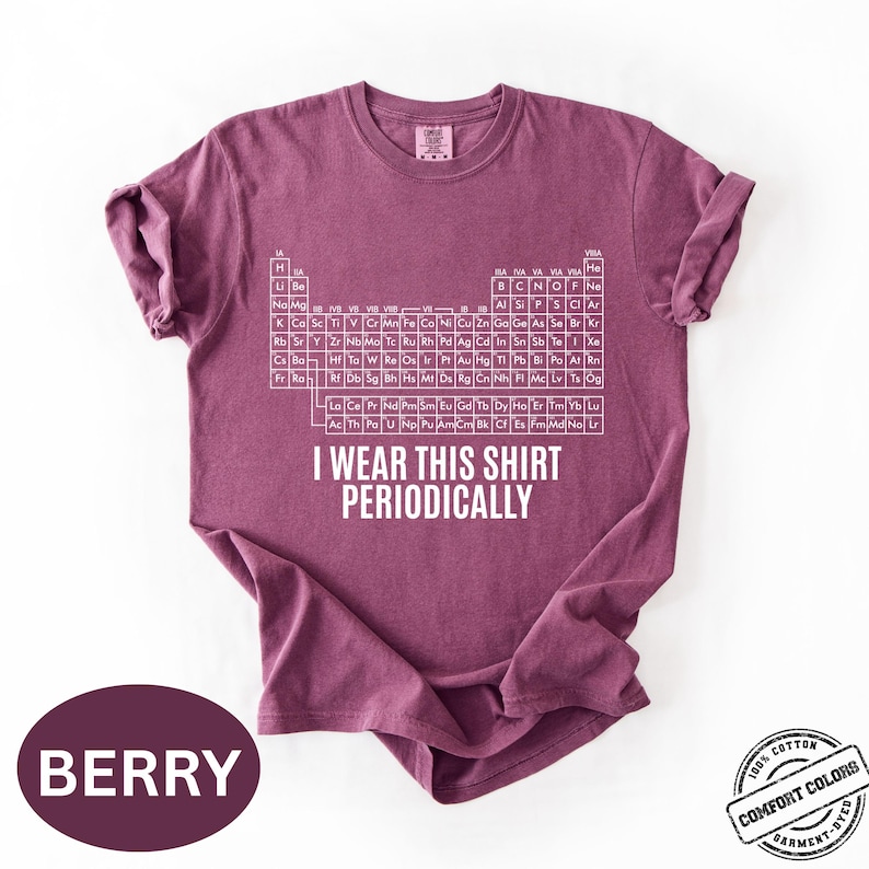 Może przedstawiać: Koszulka w kolorze jagodowym z białym wzorem układu okresowego i napisem "I WEAR THIS SHIRT PERIODICALLY". Koszulka wykonana w 100% z bawełny i farbowana w kawałku.