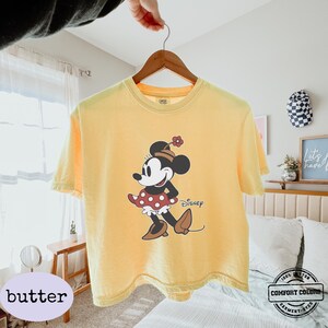 May include: A yellow t-shirt featuring a Minnie Mouse graphic. The cartoon character is depicted in a red and white polka dot dress, brown shoes, and a hat with a flower. The word "Disney" is printed below the character.