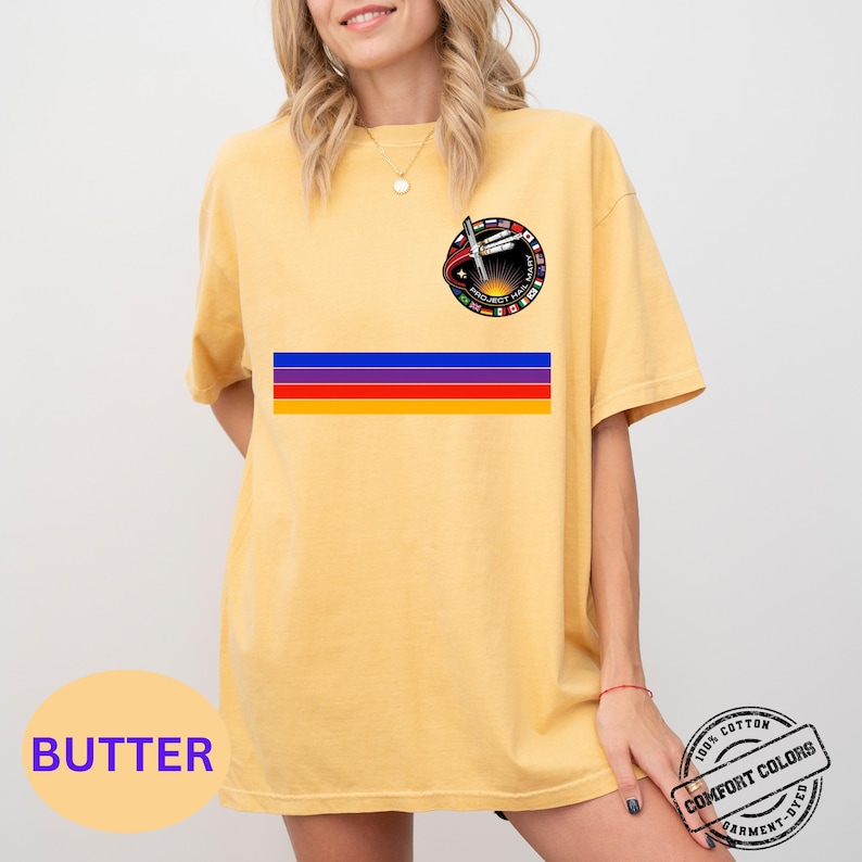 May include: Butter-colored t-shirt with a circular graphic on the upper left chest and a horizontal stripe design in blue, purple, red, and yellow. The shirt also has a Comfort Colors garment-dyed label.