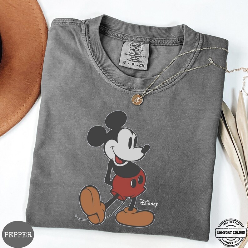 May include: A grey Comfort Colors t-shirt with a Mickey Mouse graphic in black, red, and brown. A silver necklace with a pendant is draped across the shirt. The shirt is labelled "Pepper". The t-shirt is made of 100% cotton.