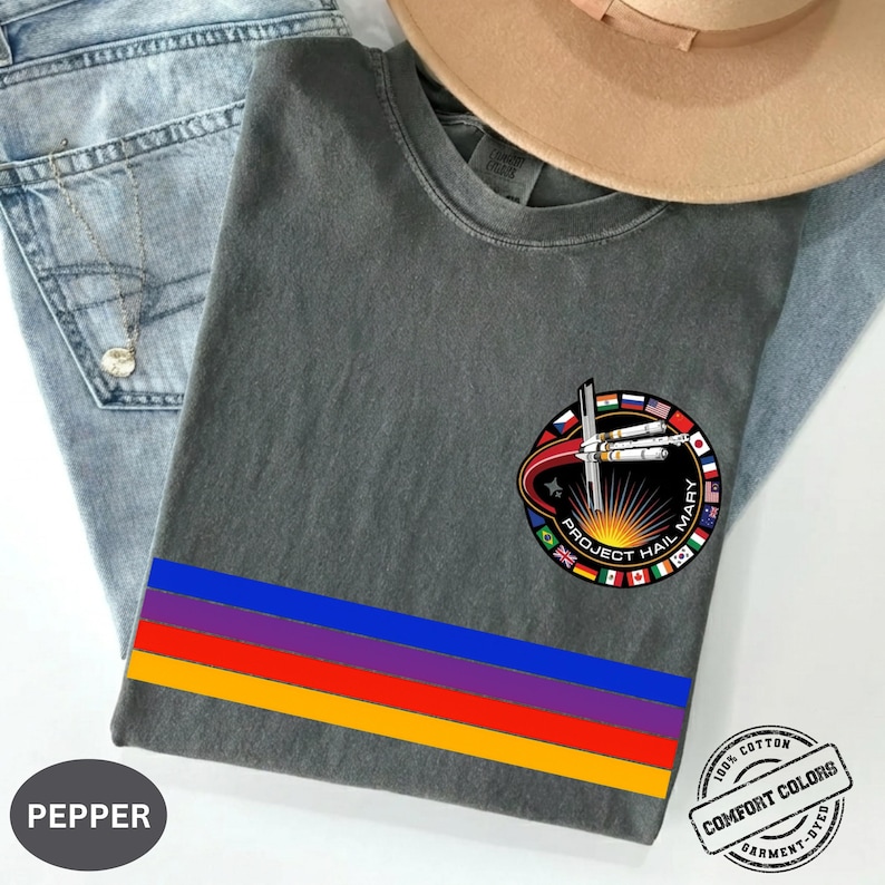 May include: A gray t-shirt with a colorful horizontal stripe design and a circular patch featuring the text "Project Hail Mary" and flags. The shirt is made of 100% cotton and is garment-dyed.