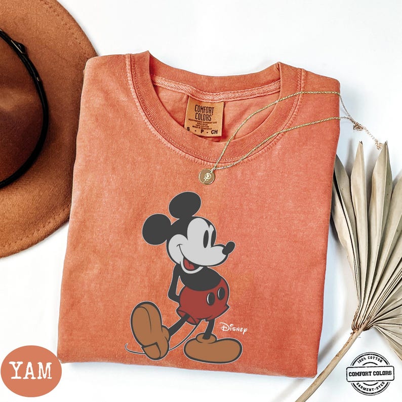 May include: An orange Comfort Colors t-shirt with a Mickey Mouse graphic. The cartoon character is in black, white, and red. A gold necklace and a brown hat are visible. The shirt has the Disney logo and the Comfort Colors logo.