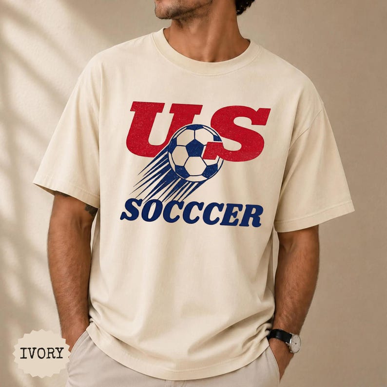 K&ouml;nnte beinhalten: Elfenbeinfarbenes T-Shirt mit rotem "US"-Schriftzug und einem Fu&szlig;ball-Design. Das Wort "SOCCER" ist in Marineblau unter dem Fu&szlig;ball aufgedruckt. Das Shirt ist ein l&auml;ssiger Unisex-Stil.