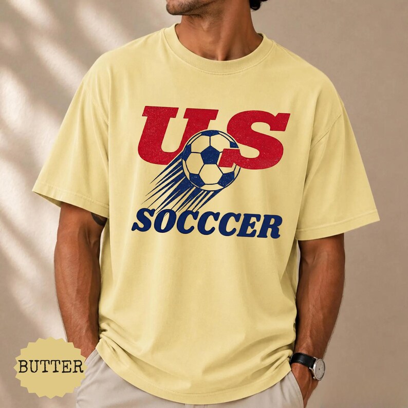K&ouml;nnte beinhalten: Hellgelbes T-Shirt mit dem Text "US SOCCER" in Rot und Blau. Das Design beinhaltet einen Fu&szlig;ball mit Geschwindigkeitslinien. Das Shirt ist ein l&auml;ssiger Stil.