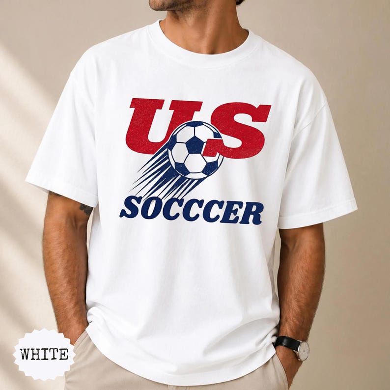 K&ouml;nnte beinhalten: Wei&szlig;es T-Shirt mit einem grafischen Design mit dem Text "US SOCCER" in Rot und Blau. Ein Fu&szlig;ball mit blauen und wei&szlig;en F&uuml;nfecken ist in das Design integriert, mit Geschwindigkeitslinien.