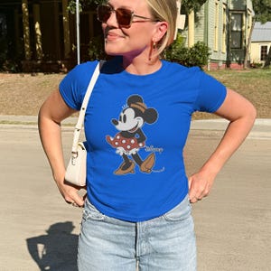 May include: Royal blue t-shirt featuring a Minnie Mouse graphic. The cartoon character is depicted in a classic pose, wearing a red polka-dot skirt, brown shoes, and a matching hat. The Disney logo is printed below the graphic.