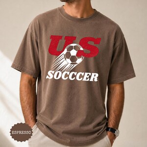 K&ouml;nnte beinhalten: Espressofarbenes T-Shirt mit dem Text "US SOCCER" in Rot und Wei&szlig;. Das Design zeigt einen Fu&szlig;ball mit Bewegungslinien. Das Shirt ist kurz&auml;rmlig und hat einen Rundhalsausschnitt.