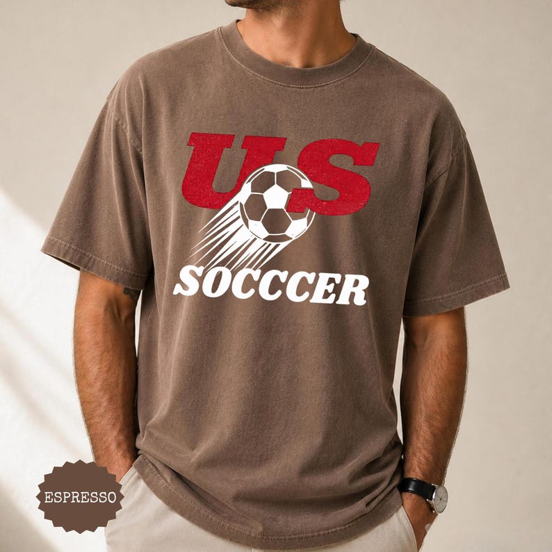 K&ouml;nnte beinhalten: Espressofarbenes T-Shirt mit dem Text "US SOCCER" in Rot und Wei&szlig;. Das Design zeigt einen Fu&szlig;ball mit Bewegungslinien. Das Shirt ist kurz&auml;rmlig und hat einen Rundhalsausschnitt.