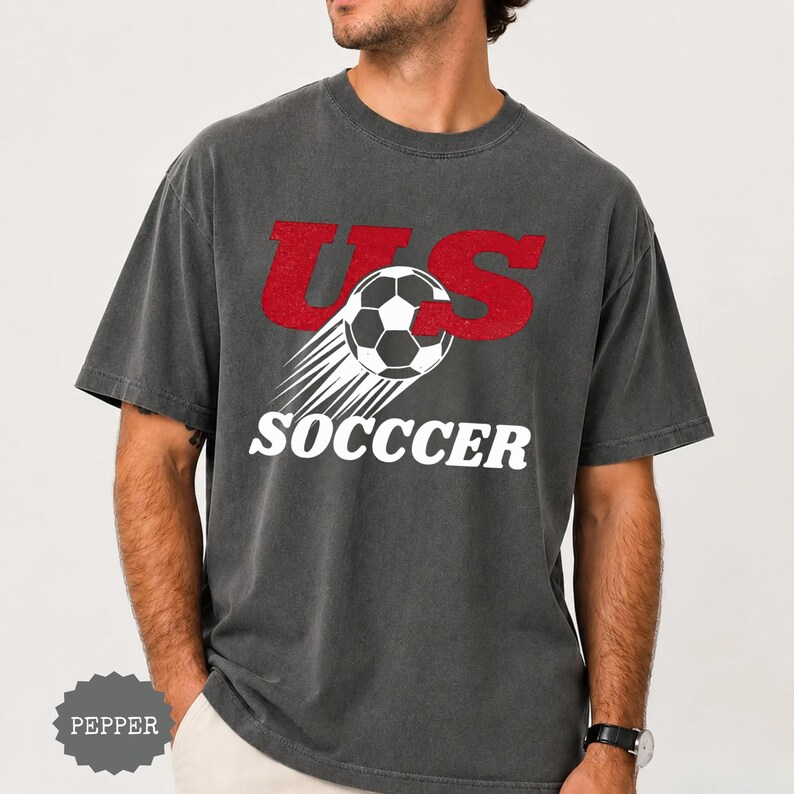 K&ouml;nnte beinhalten: Dunkelgraues T-Shirt mit dem Text "US SOCCER" in Rot und Wei&szlig;. Das Design zeigt einen Fu&szlig;ball mit Geschwindigkeitslinien. Das Shirt hat einen Vintage-Stil.
