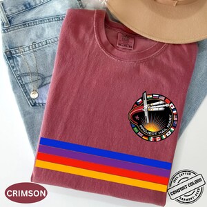 May include: Crimson-colored t-shirt with a circular graphic featuring the text "PROJECT HAIL MARY" and a design of a rocket and flags. The shirt has a horizontal stripe design in blue, red, purple, and yellow. The shirt is made of 100% cotton.