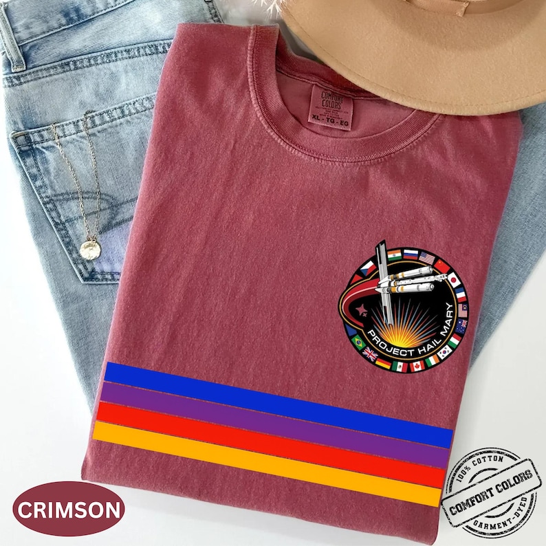 May include: Crimson-colored t-shirt with a circular graphic featuring the text "PROJECT HAIL MARY" and a design of a rocket and flags. The shirt has a horizontal stripe design in blue, red, purple, and yellow. The shirt is made of 100% cotton.