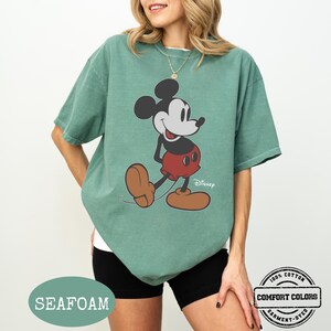 May include: Seafoam green t-shirt with a Mickey Mouse graphic. The cartoon character is in black, red, and brown. The shirt is labelled "Comfort Colors" and is made of 100% cotton.