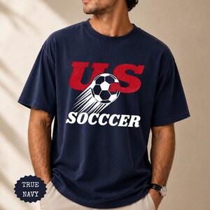 K&ouml;nnte beinhalten: Marineblaues T-Shirt mit dem Text "US SOCCER" in Rot und Wei&szlig;. Ein Fu&szlig;ball mit Bewegungslinien ist im Design enthalten. Das T-Shirt hat die Farbe True Navy.