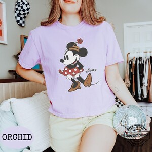May include: A light purple t-shirt featuring a classic Minnie Mouse graphic with the word "Disney". The shirt is a Comfort Colors brand and is made of 100% cotton. The word "ORCHID" is printed at the bottom.