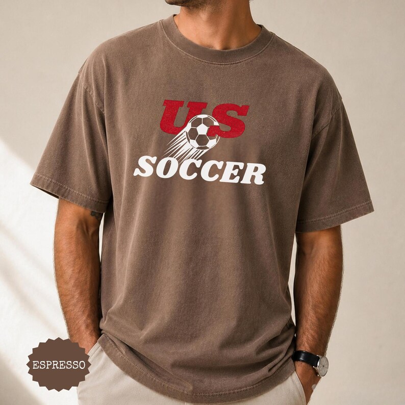 Op de afbeelding: Espresso-kleurig t-shirt met de tekst "US SOCCER" in rood en wit. Het ontwerp bevat een voetbal met bewegingslijnen. Het shirt heeft korte mouwen en een ronde hals.