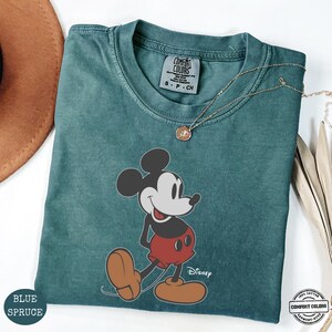 May include: A blue spruce Comfort Colors t-shirt with a Mickey Mouse graphic in red shorts and brown shoes. The Disney logo is printed below the character. A gold necklace is draped across the shirt.