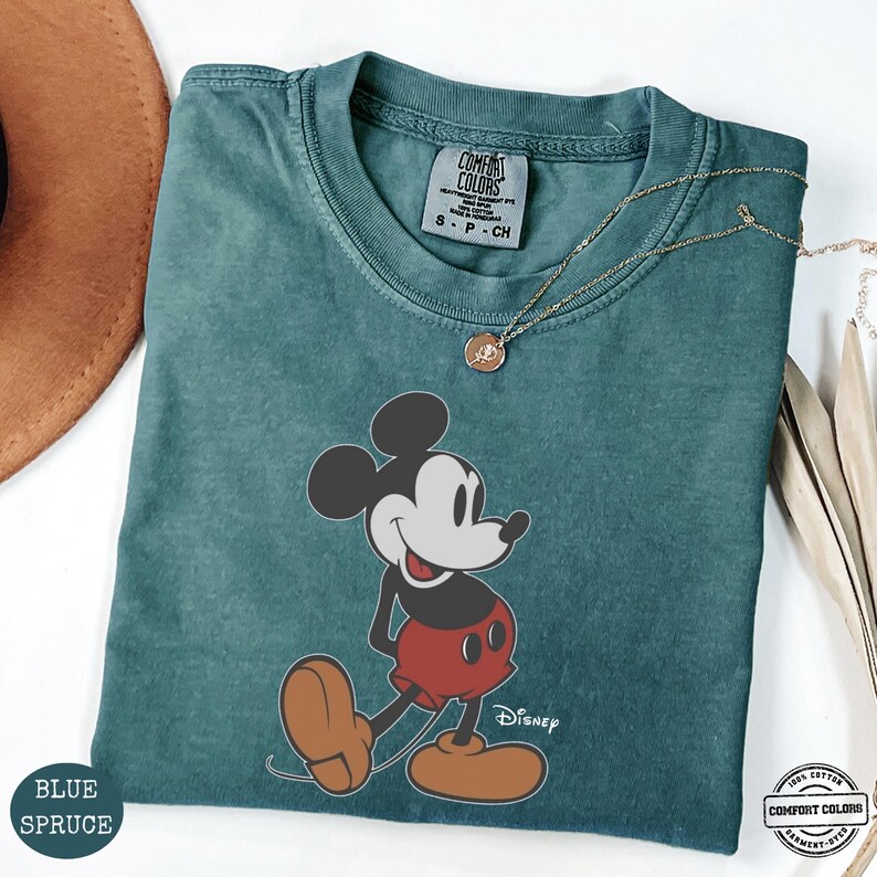 May include: A blue spruce Comfort Colors t-shirt with a Mickey Mouse graphic in red shorts and brown shoes. The Disney logo is printed below the character. A gold necklace is draped across the shirt.