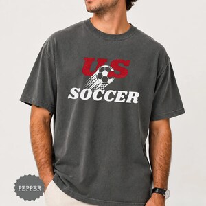 Op de afbeelding: Donkergrijs t-shirt met de tekst "US Soccer" in rood en wit. Het ontwerp bevat een voetbal met bewegingslijnen. Het shirt heeft een casual stijl.