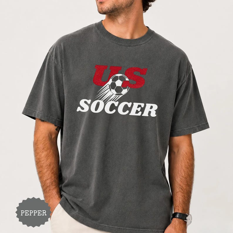 Op de afbeelding: Donkergrijs t-shirt met de tekst "US Soccer" in rood en wit. Het ontwerp bevat een voetbal met bewegingslijnen. Het shirt heeft een casual stijl.