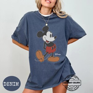 May include: Denim blue t-shirt with a Mickey Mouse graphic. The cartoon character is shown in red shorts, brown shoes, and black ears. The shirt has the text "Disney" and "Comfort Colors" printed on it.