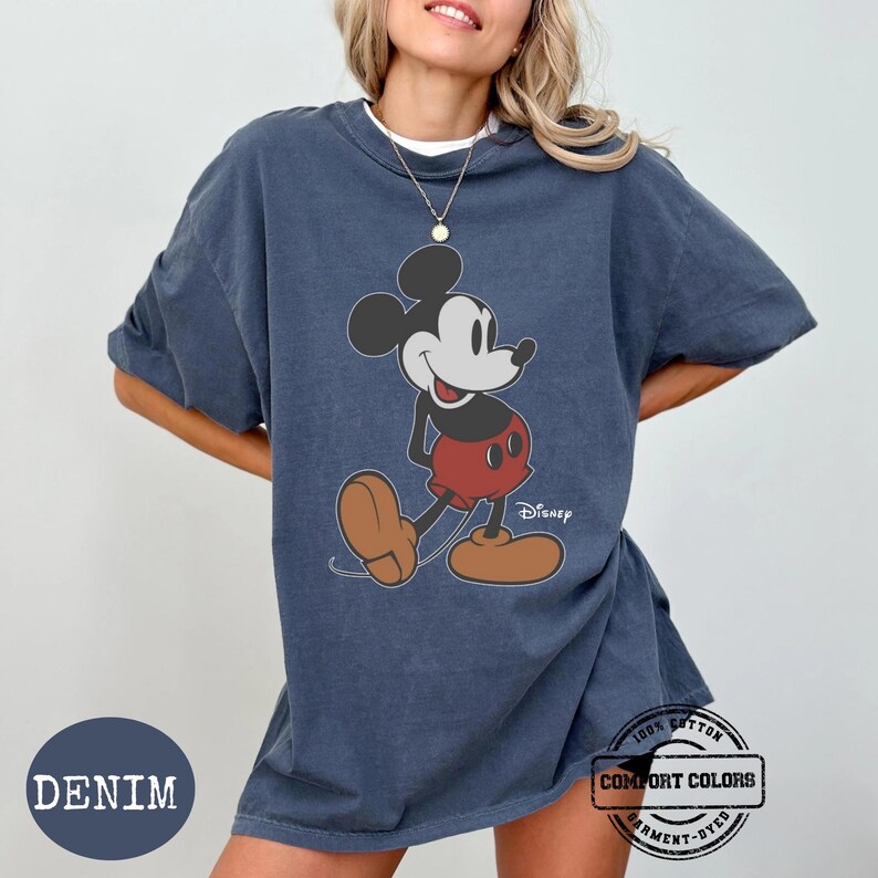May include: Denim blue t-shirt with a Mickey Mouse graphic. The cartoon character is shown in red shorts, brown shoes, and black ears. The shirt has the text "Disney" and "Comfort Colors" printed on it.