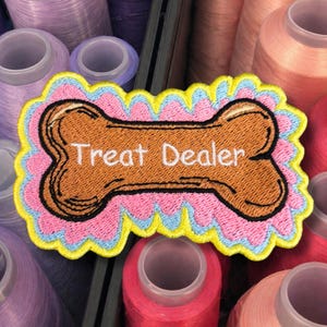 May include: Embroidered patch in the shape of a dog bone with the words "Treat Dealer" in white. The patch has a brown bone design, a pink background, blue and yellow trim, and a black outline. The background includes spools of thread.
