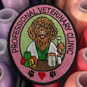 May include: Embroidered patch with the text "PROFESSIONAL VETERINARY CLINIC" around a cartoon dog wearing a lab coat. The patch features a green and pink colour scheme, with paw prints and a red heart at the bottom. The background includes spools of thread.