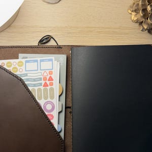 Peut inclure: Un journal en cuir marron avec une couverture noire et une poche contenant des autocollants. Les autocollants ont diverses formes et couleurs, notamment des rectangles bleus, des triangles rouges et des cercles jaunes. Le journal est ouvert, révélant les pages intérieures.