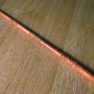 Puede incluir: Una varita larga y delgada de madera de color marrón claro con intrincados patrones geométricos tallados a lo largo del eje. La varita se estrecha hasta una punta fina en un extremo y mantiene un acabado suave y pulido en toda su longitud.