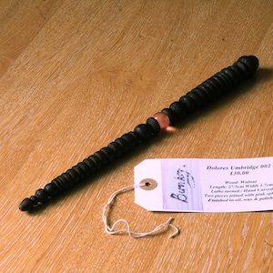 Puede incluir: Una varita de madera oscura con un diseño de cuentas, con una esfera rosa en el medio. La varita mide aproximadamente 27,5 cm de largo. Una etiqueta con el texto "Dolores Umbridge 002" y otros detalles está adjunta.