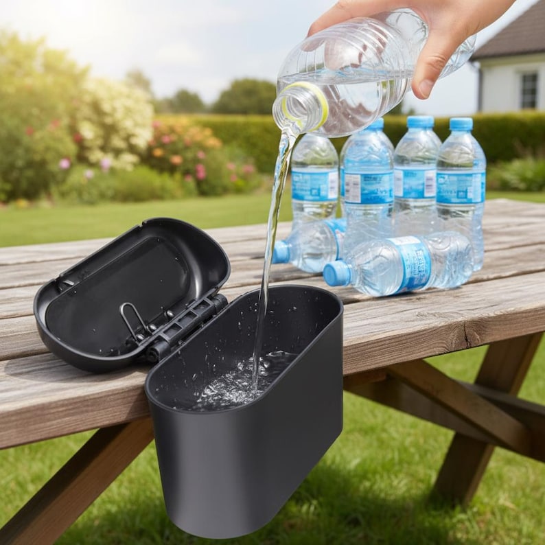 May include: A black, rectangular container with a hinged lid is open on a wooden picnic table. Water is being poured from a clear plastic bottle into the container. Several water bottles are in the background, with a green lawn and trees.