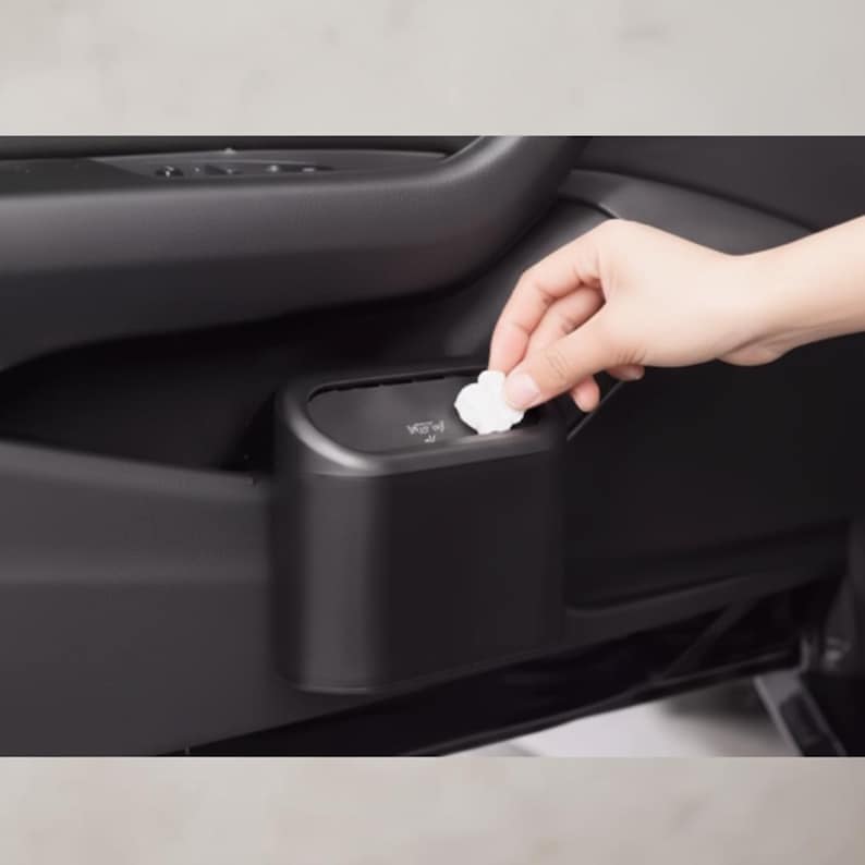 May include: A black car trash bin installed in a car door. A hand is placing a white tissue into the bin. The bin is rectangular with a slightly curved top. The text "Vow" is visible on the bin.