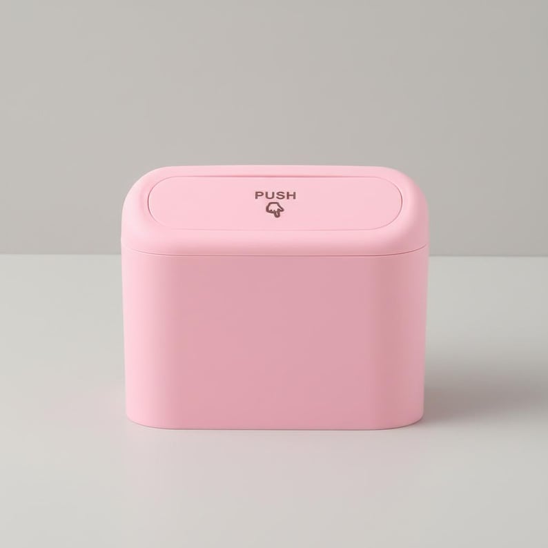May include: A small, rectangular, pink trash can with a rounded top. The lid is labeled "PUSH" with a small graphic. The container is a solid, pastel pink color and sits on a light gray surface. The design is simple and modern.