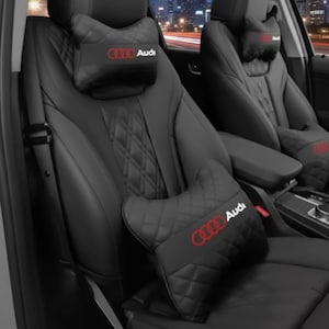 Puede incluir: Cojines negros para reposacabezas de coche con el logotipo rojo de Audi. Los cojines tienen un diseño acolchado de diamantes y están colocados en asientos de coche de cuero negro. Se ve el interior del coche.