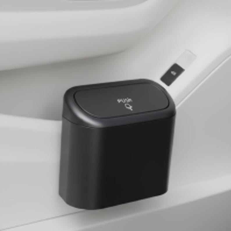 May include: A small, black, rectangular trash can with a rounded top. The lid is labeled "Push" with a small arrow. The trash can is mounted on a white surface, likely in a vehicle.