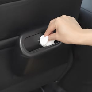 May include: A hand placing a white wipe into a black car interior compartment. The compartment has a curved design and the letter "H" is visible. The car interior is black.