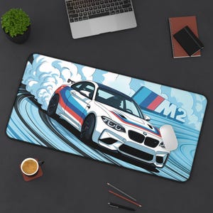 May include: White mousepad featuring a stylized illustration of a white sports car with blue, red, and white racing stripes, drifting on a blue track. The word "M2" is displayed in blue. A computer mouse is visible.
