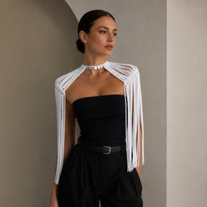 May include: A white macrame shoulder wrap featuring long, dangling fringe and a choker-style neckline adorned with small, circular silver-toned metal discs. The piece is constructed from knotted cord and rests over the shoulders and upper arms.